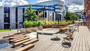 A modern building with a curved glass facade, outdoor seating area with wooden benches and tables, and a wooden deck with steps leading to a water feature.