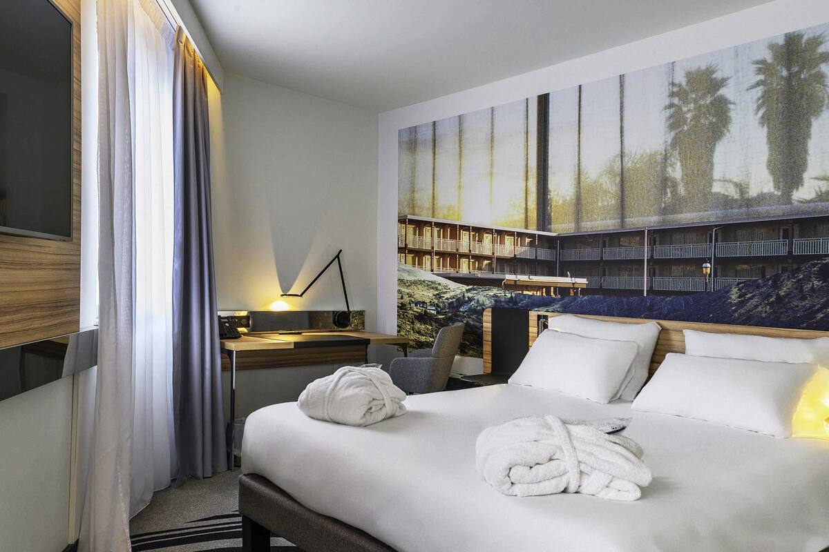 A hotel room with a bed, a desk with a lamp, a large painting on the wall, and a window with curtains.