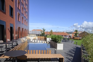 An outdoor terrace with wooden tables and chairs, a pool, and a view of buildings in the background. - Novotel Saint-Étienne Centre Gare Châteaucreux (Saint-Etienne)