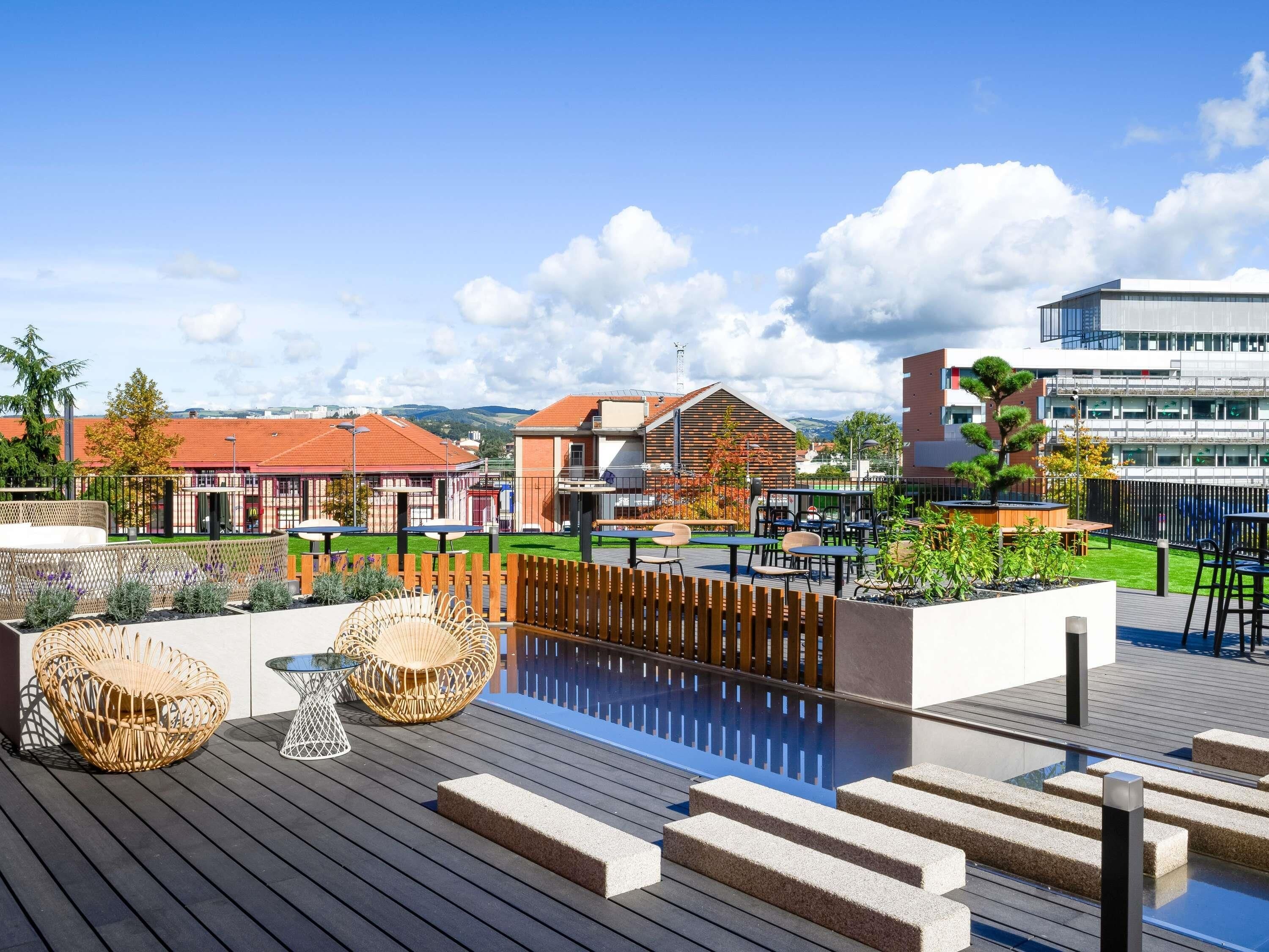 A rooftop pool area with wooden decking, lounge chairs, and a view of buildings in the background.