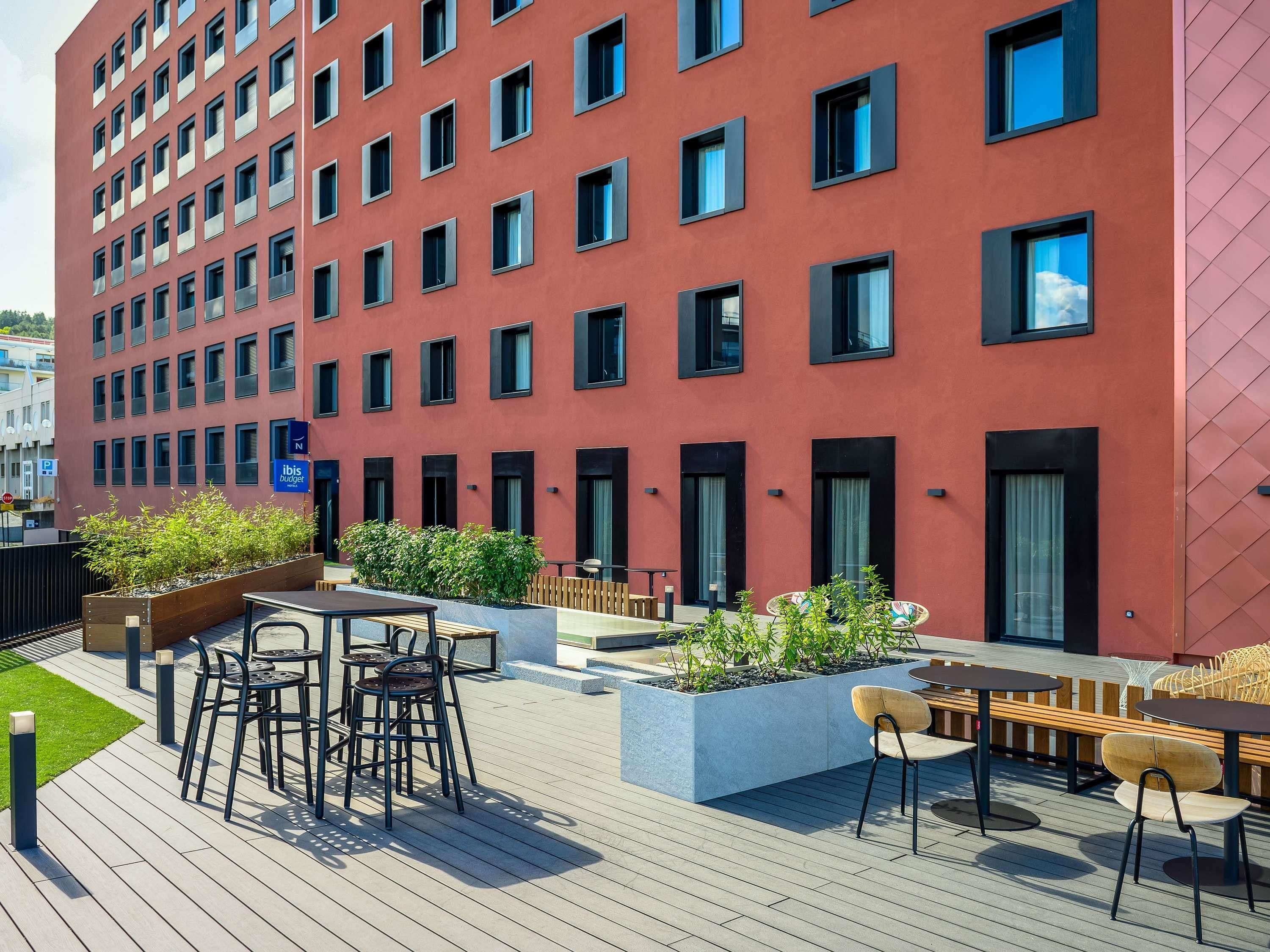 Un bâtiment moderne avec une façade rouge, de nombreuses fenêtres, un espace extérieur aménagé avec des tables et des chaises, et une enseigne indiquant « ibis budget ».