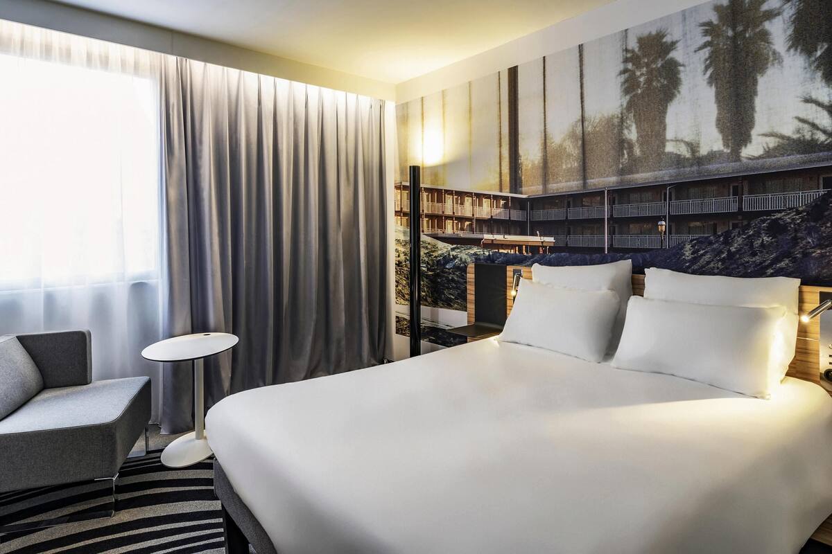 A hotel room with a large bed, a desk, a chair, and a view of a building with balconies.