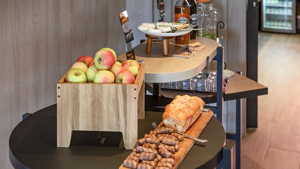 A wooden fruit bowl with apples, a tray of chocolate-covered snacks, and a loaf of bread on a round table. - Novotel Saint-Étienne Centre Gare Châteaucreux (Saint-Etienne)
