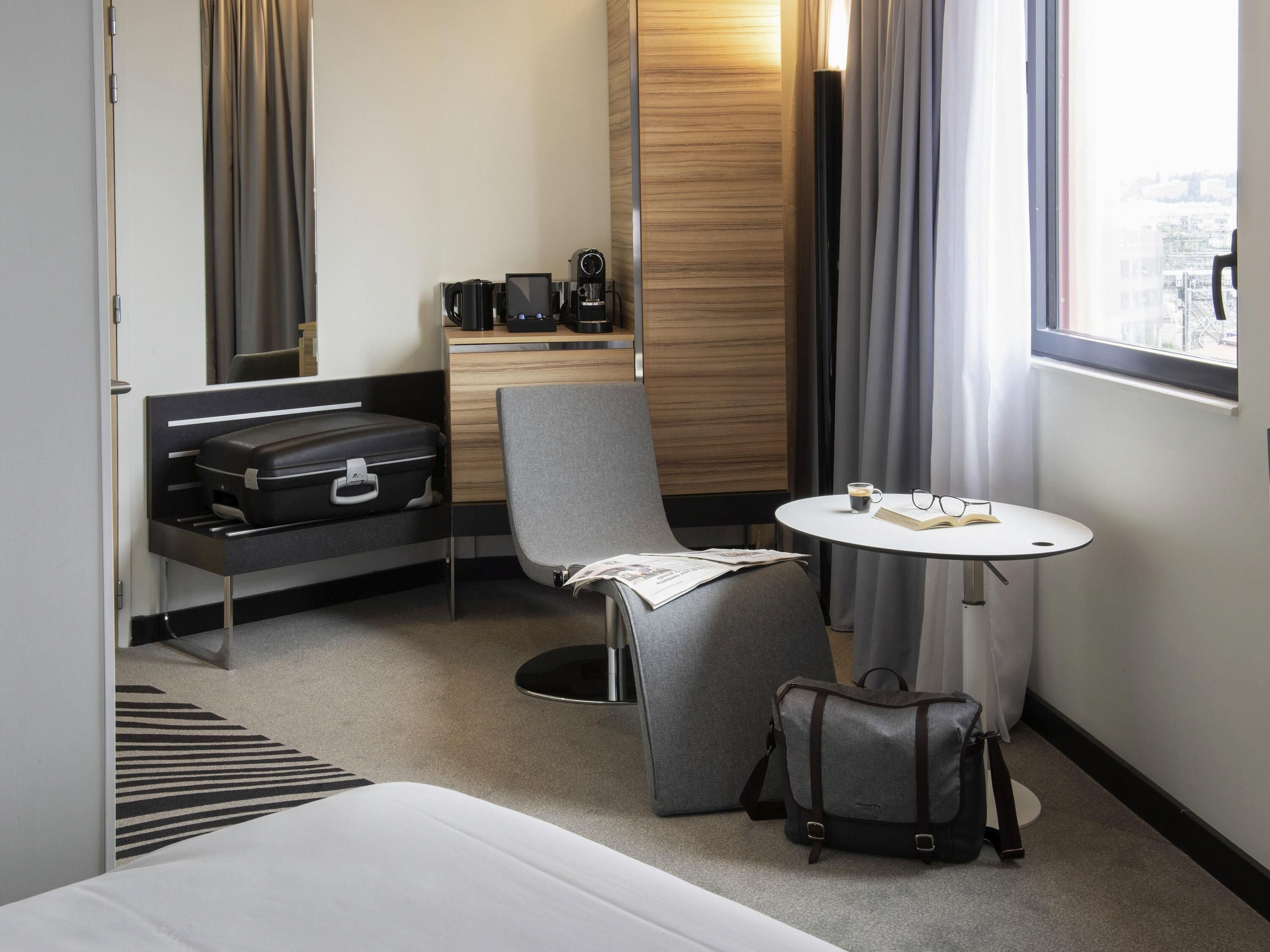 Een hotelkamer met een bed, een bureau met koffiezetapparaat, een stoel, een tafel met een boek en brillen, en een koffer.