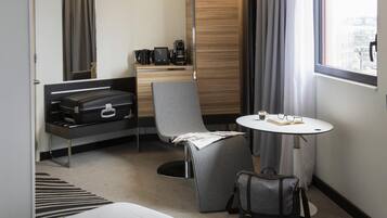Een hotelkamer met een bed, een bureau met koffiezetapparaat, een stoel, een tafel met een boek en brillen, en een koffer.
