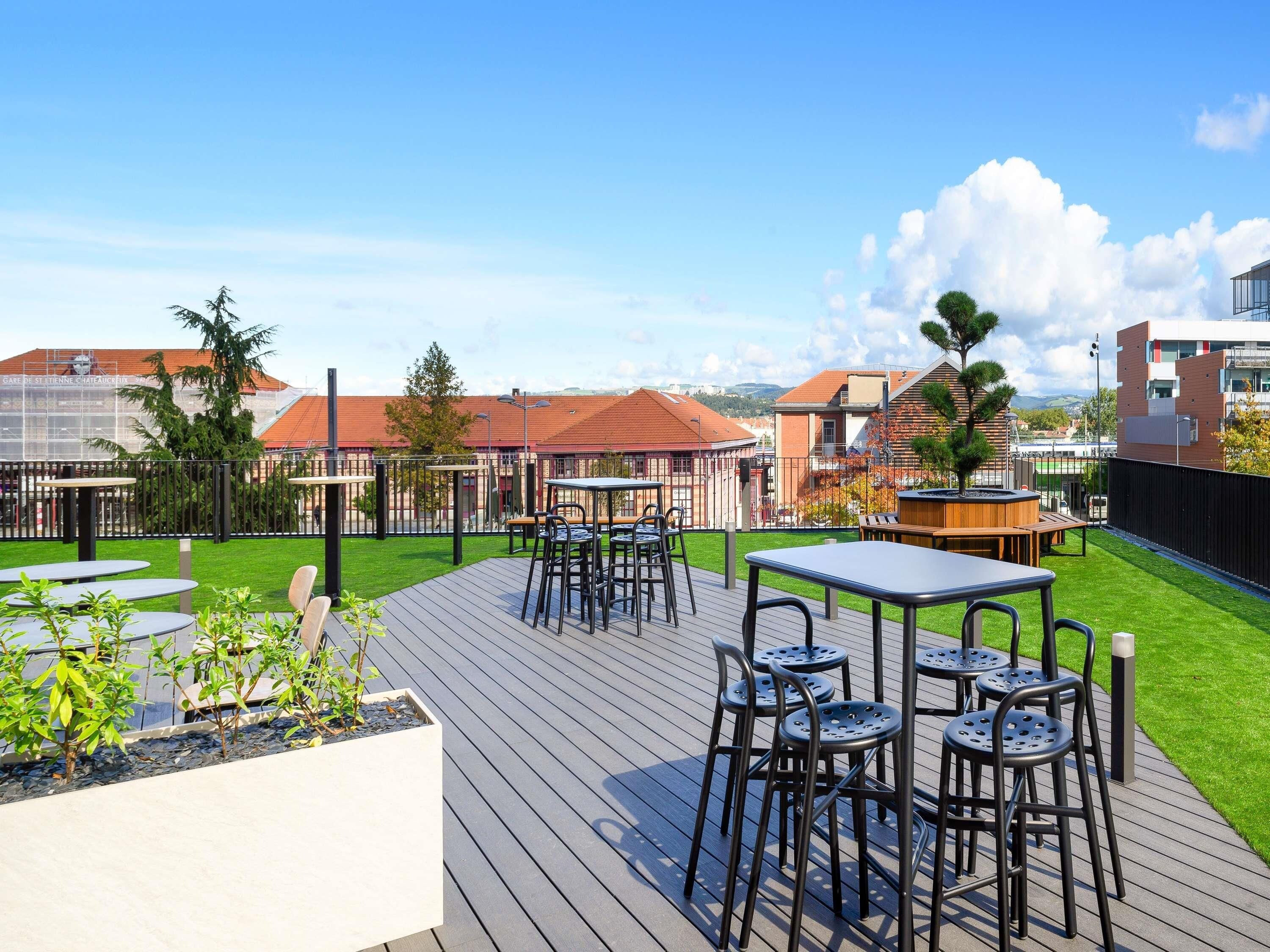 Une terrasse extérieure avec des tables et des chaises, une plateforme en bois et une vue sur les bâtiments en arrière-plan.