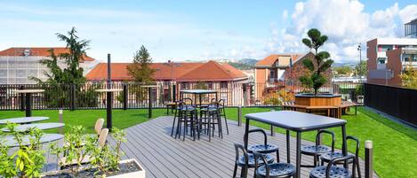 Une terrasse extérieure avec des tables et des chaises, une plateforme en bois et une vue sur les bâtiments en arrière-plan.