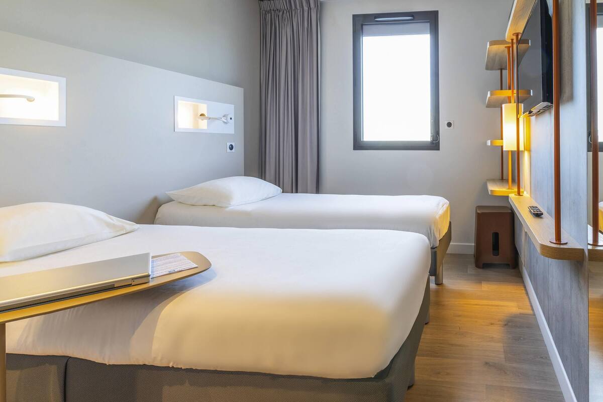 A hotel room with two beds, a window, a bedside table with a lamp, and a small stool.