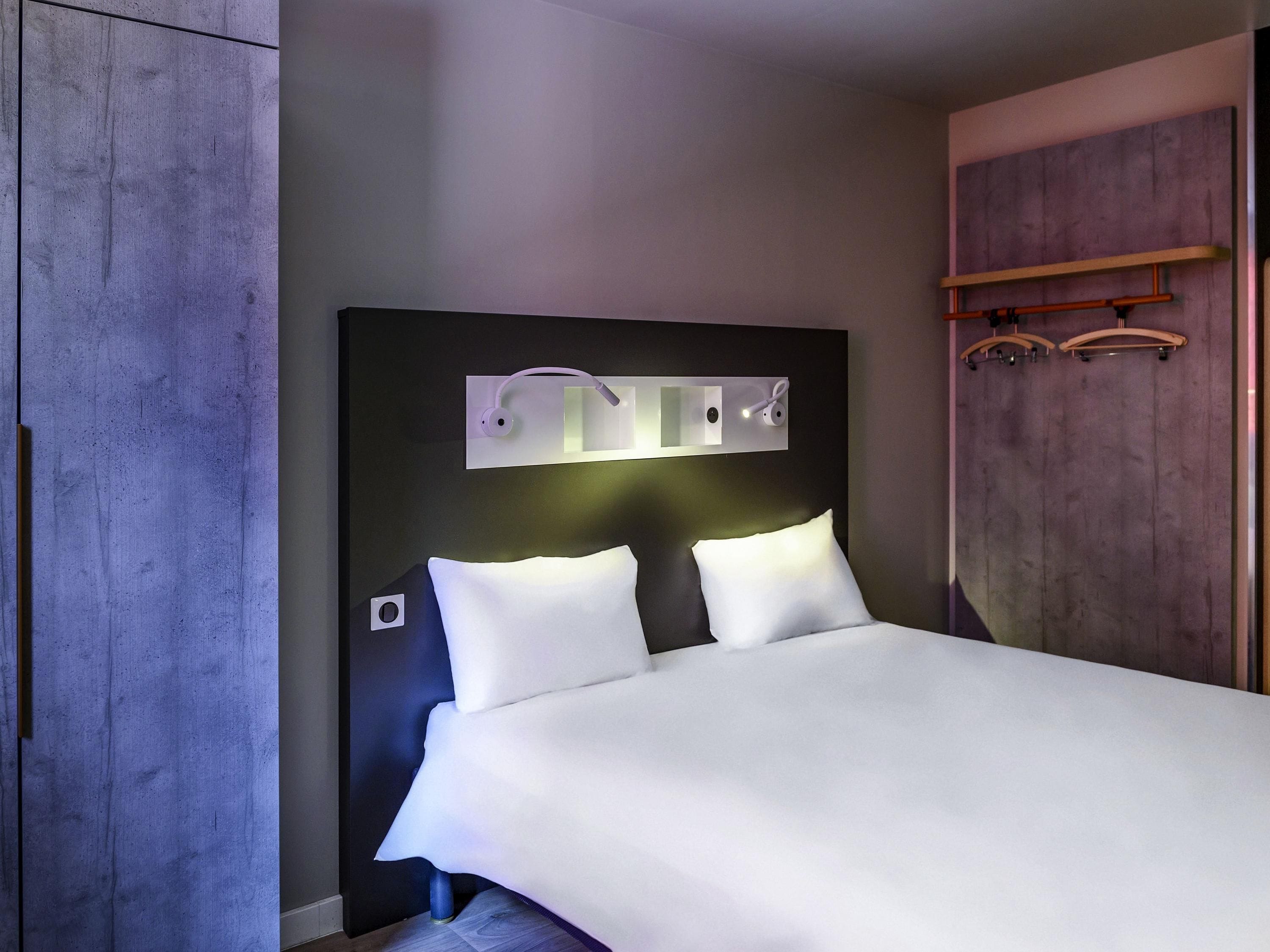 Une chambre d’hôtel moderne dotée d’un grand lit, d’une tête de lit avec éclairage intégré, d’une armoire et d’un porte-manteau.