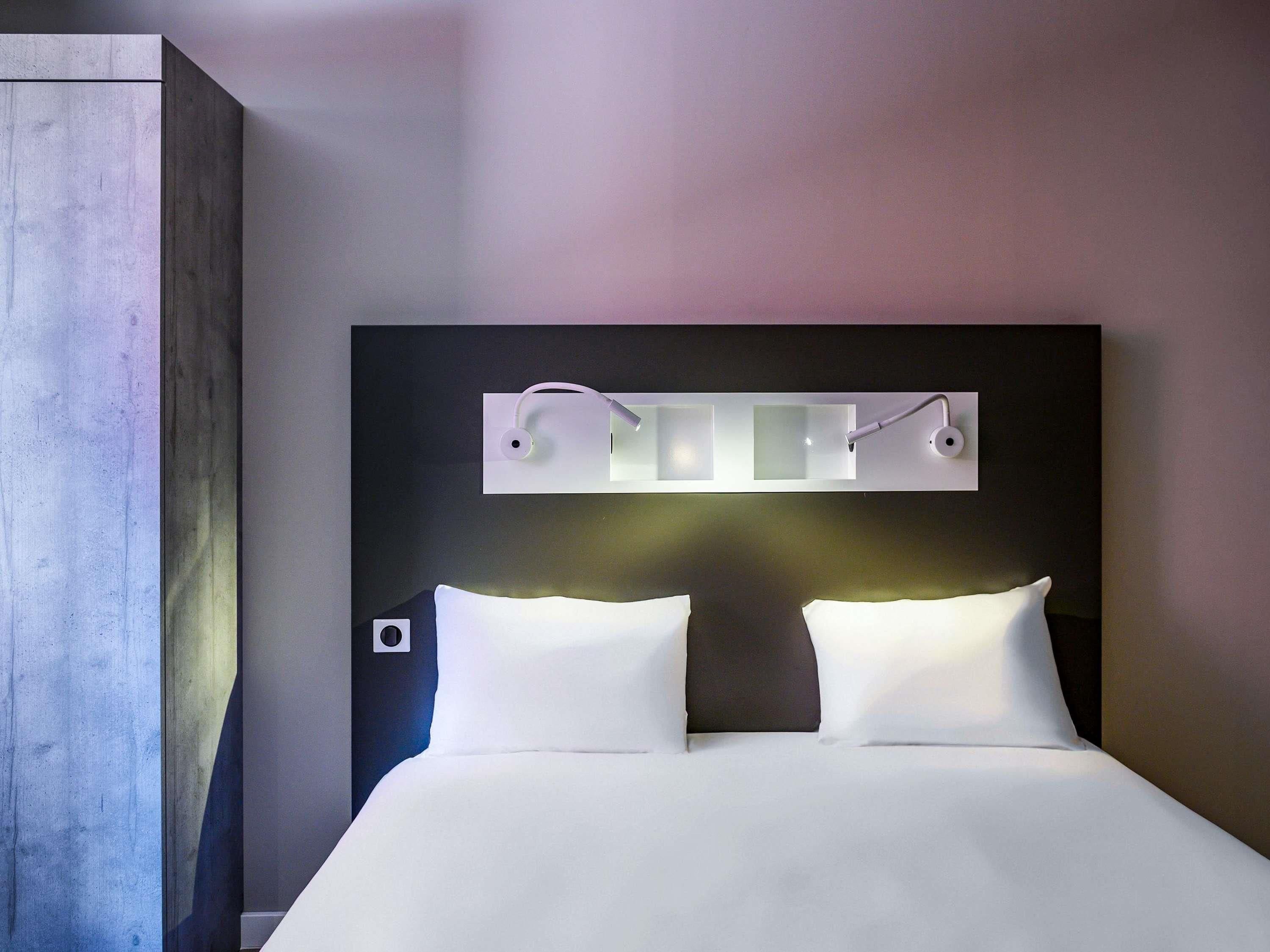 A hotel room with a bed, a headboard with built-in lighting, and a wooden wardrobe.