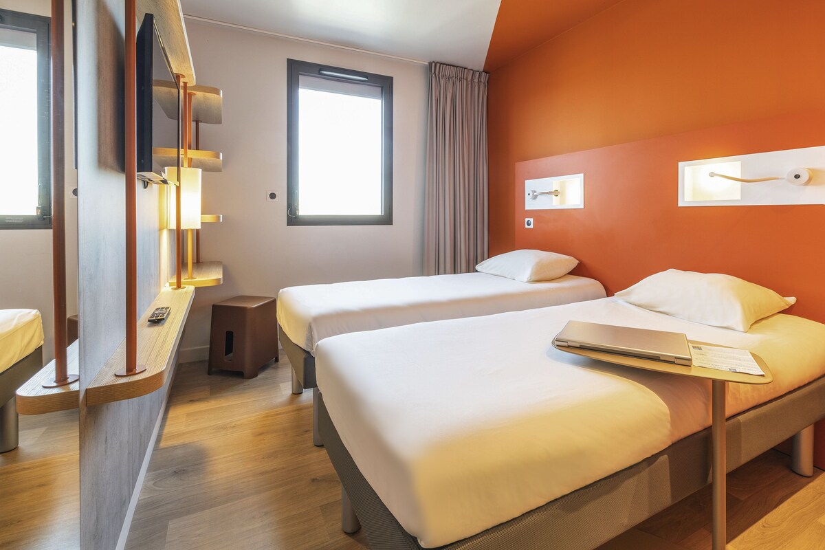 Une chambre d’hôtel avec deux lits, un bureau avec un ordinateur portable, une fenêtre et un rideau.