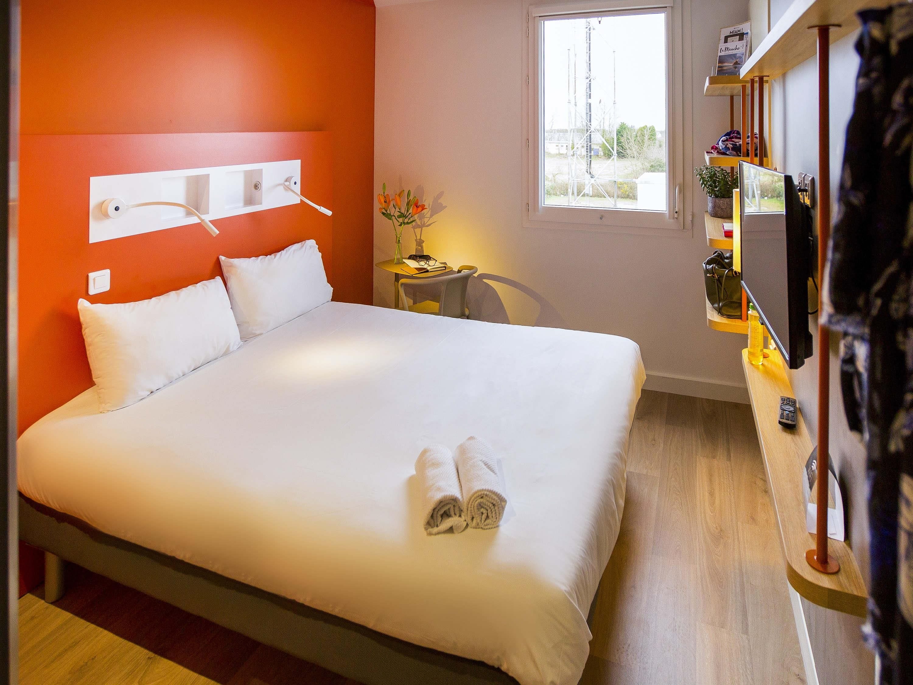 A hotel room with a large bed, a small table with a chair, a TV, and a window with a view of outside structures.