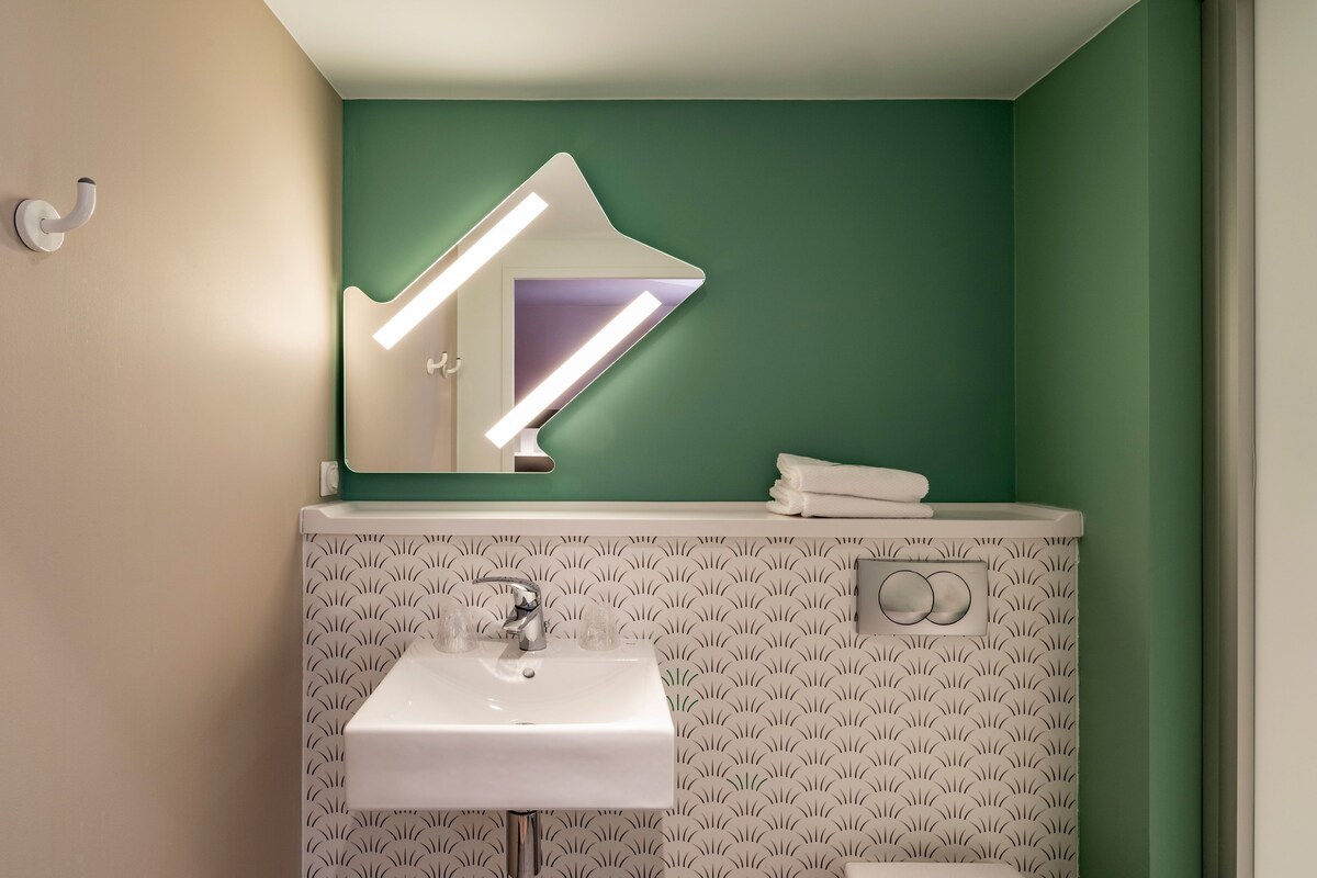 Une salle de bain avec un lavabo blanc, un miroir agrémenté d’un luminaire aux formes géométriques et un mur vert.