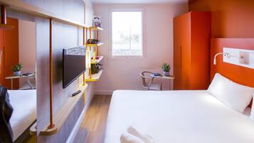 Et moderne hotelværelse med seng, fjernsyn, et lille bord med stol, et vindue og en hylde med personlige ejendele.
