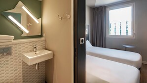 A hotel room with a bathroom featuring a sink, mirror, and towel rack. The room has two beds and a window with a view of buildings. - Ibis Budget Saint-Etienne Centre Gare Châteaucreux (Saint-Etienne)