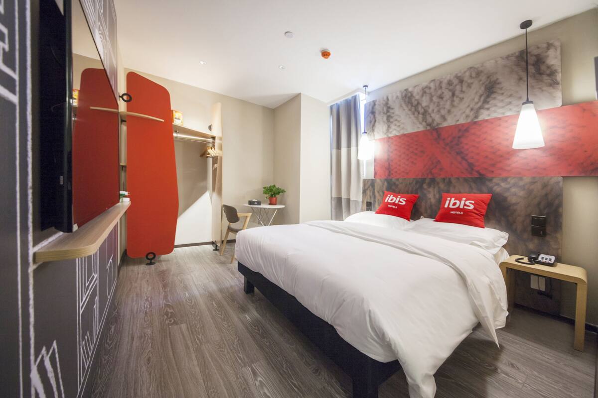 A modern hotel room with a large bed, a desk, and a chair. There is a red wall with the word "ibis" on it.