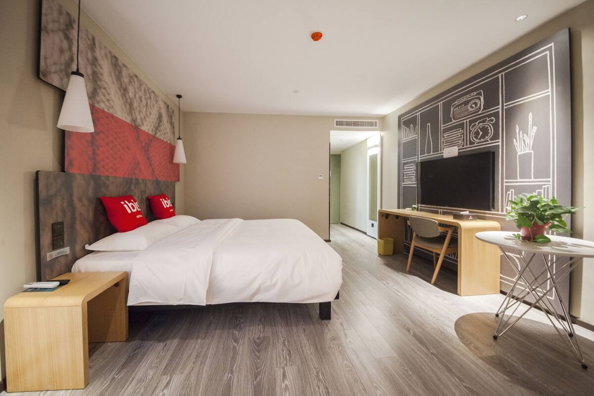 A modern hotel room with a large bed, bedside table, and a desk with a chair.