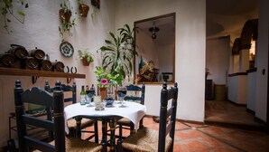 Un comedor con una mesa redonda, sillas y un mantel blanco. Hay plantas en macetas en las paredes y un suelo de baldosas.