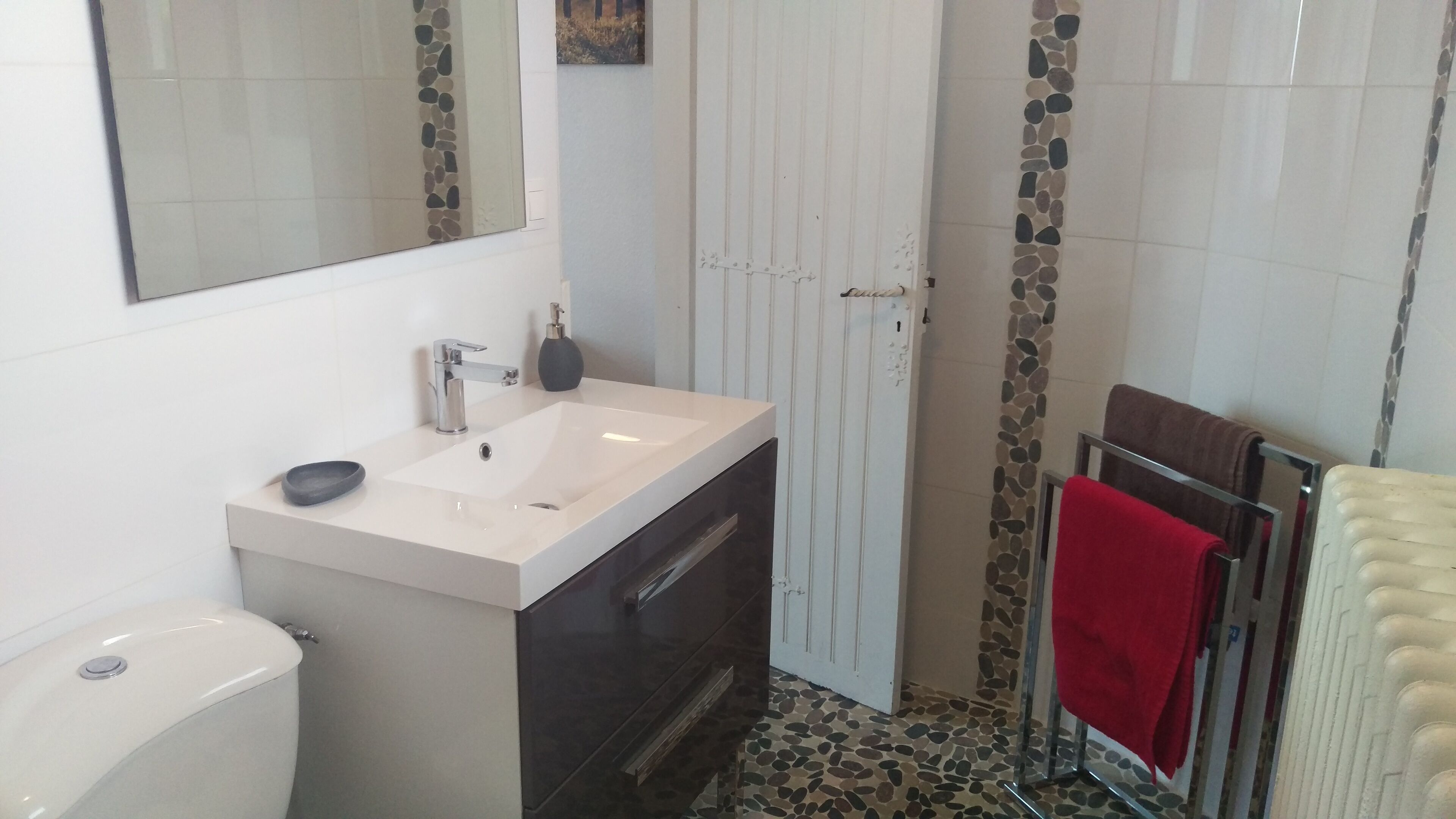 A bathroom with a white sink, dark cabinet, and a mirror above it. There is a towel rack with a red towel and a beige towel. The floor has a pebble pattern.