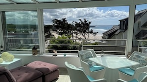 En solig balkong med utsikt över stranden, möblerad med ett vitt bord och stolar samt en rosa soffa.
