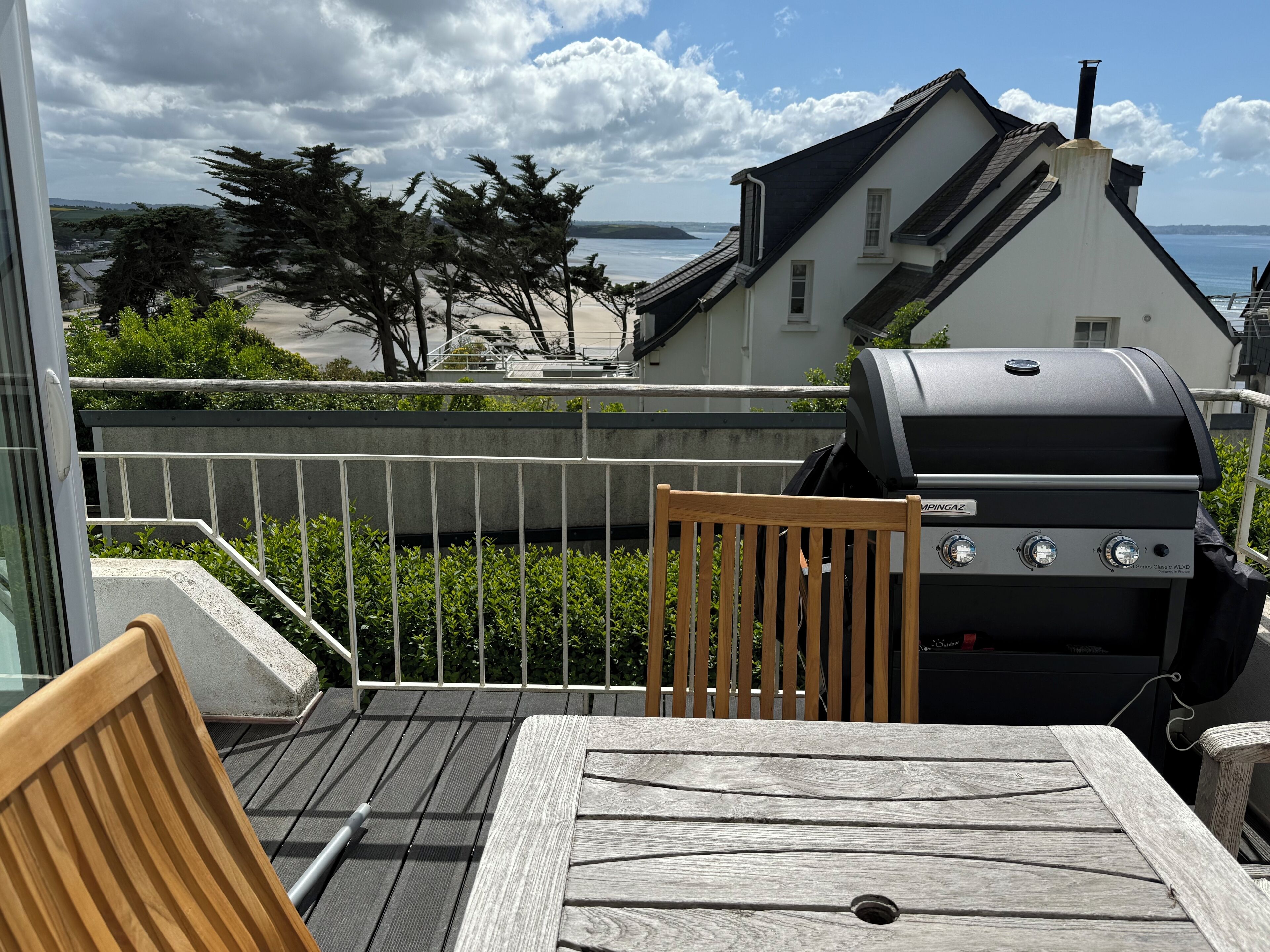 Ein Balkon mit Holztisch und -stühlen, einem Grill und Blick auf das Meer und Häuser.