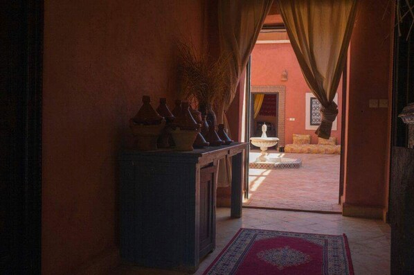 Un couloir agrémenté d’un tapis traditionnel, une table ornée de poteries et une vue sur une cour extérieure avec une fontaine.