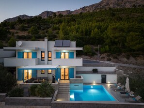 A modern two-story house with a swimming pool, outdoor seating, and a view of a mountainous landscape.