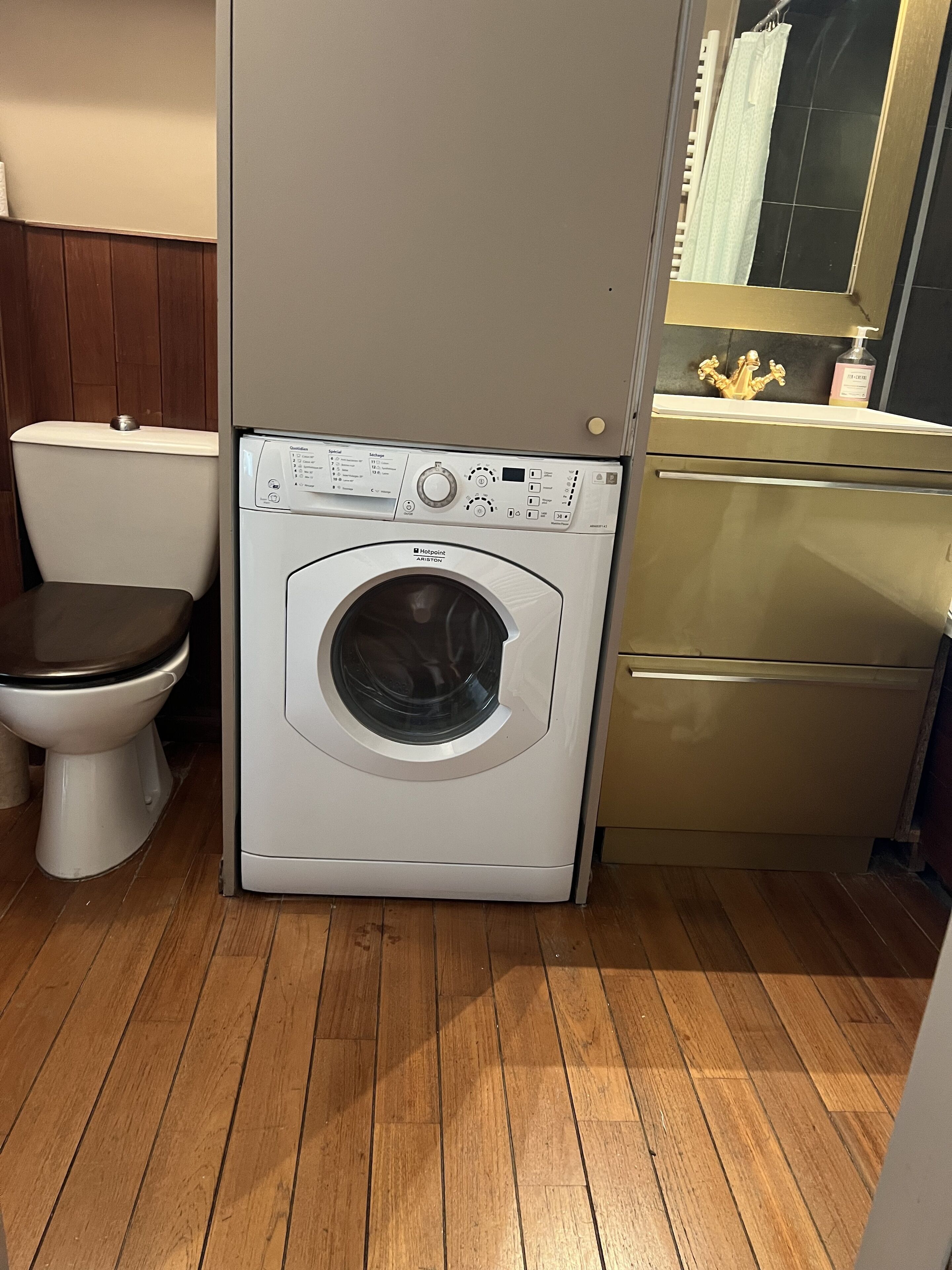 Une petite salle de bain avec un lave-linge blanc, des toilettes et un meuble de couleur dorée.