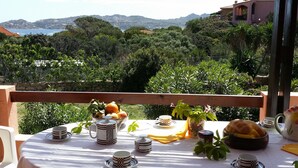 Une table dressée avec des tasses à carreaux, une théière, un pichet et un panier contenant du pain et des fruits, offrant une vue panoramique sur les montagnes et la mer.