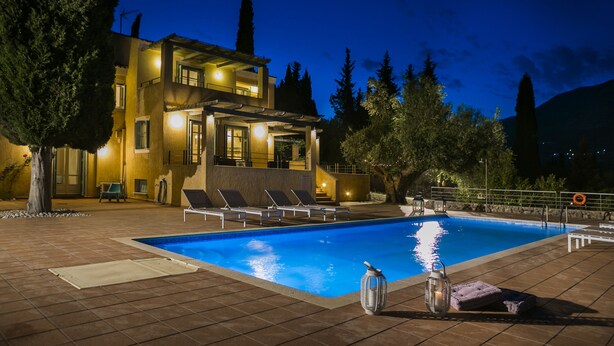 A swimming pool with lounge chairs, a pergola, and a well-lit building in the background. - 'Phivos Villa'Elegant Villa With Dramatic Mountain & Sea Views, Totally Equipped (Kefalonia)