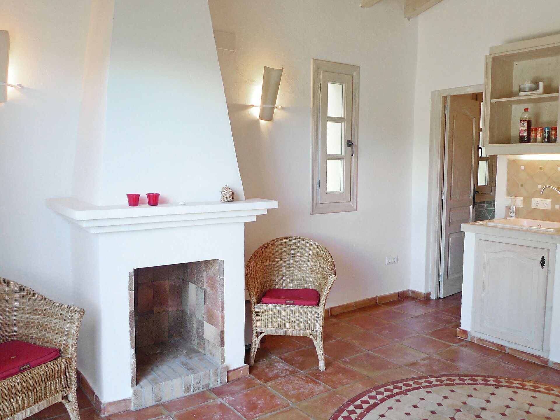 A room with a fireplace, wicker chairs, a tiled floor, and a kitchen area.