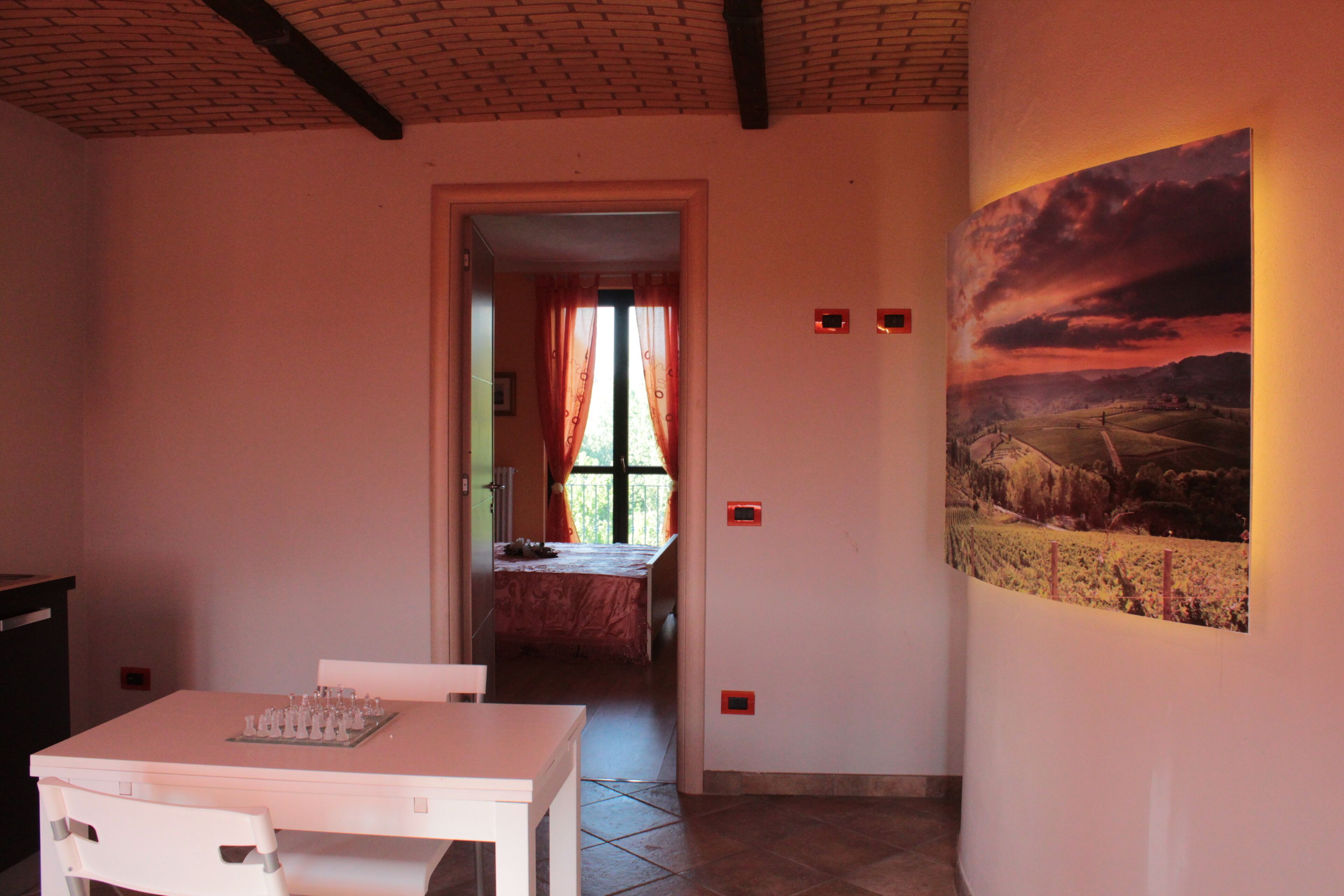 Una habitación con una mesa de comedor y sillas, una cama con cortina roja, y un cuadro de paisaje en la pared.