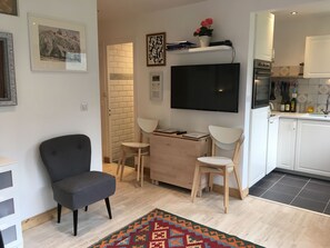 A modern living room with a grey armchair, a wooden coffee table, and a TV mounted on the wall. There is a kitchen area with white cabinets and a tiled backsplash.