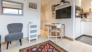A modern living room with a grey armchair, a white storage unit, a wooden desk, and a colorful rug. A flat-screen TV is mounted on the wall, and a kitchen area is visible in the background. - Le Pied du Brevent -  a studio that sleeps 4 guests (Chamonix)