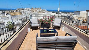 Una terraza en la azotea con mesa y sillas, una maceta con flores y vistas a la ciudad y al mar.