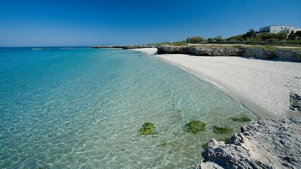 Una playa con agua turquesa cristalina, arena blanca y un promontorio rocoso.