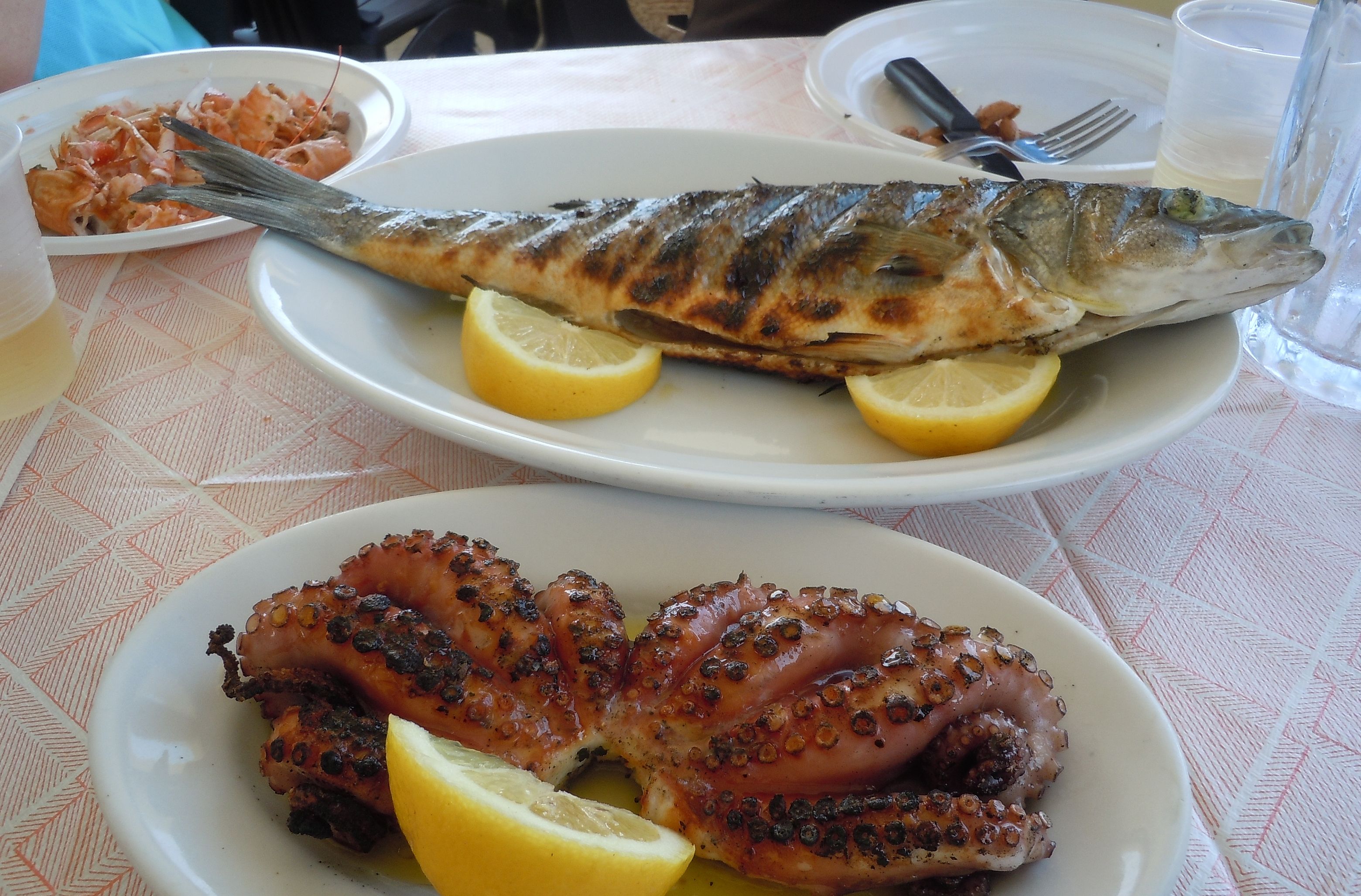 A dining table with plates of grilled fish and octopus, lemon wedges, and a fork.