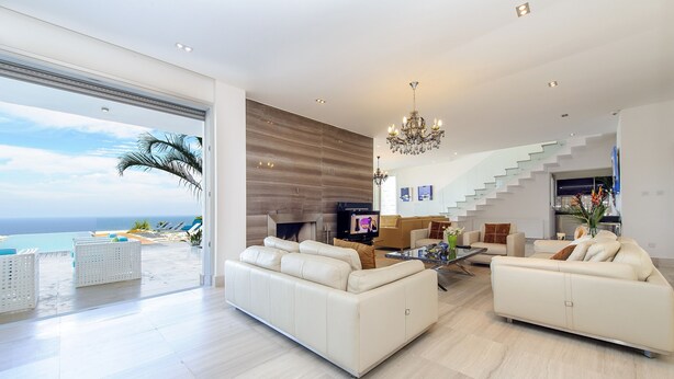 A spacious living room with a large white sofa, a coffee table, and a TV. There is a staircase leading to an upper level, and a view of the ocean through large windows. - Bay View Villa Cyprus, Luxury at its finest!! (Protaras)