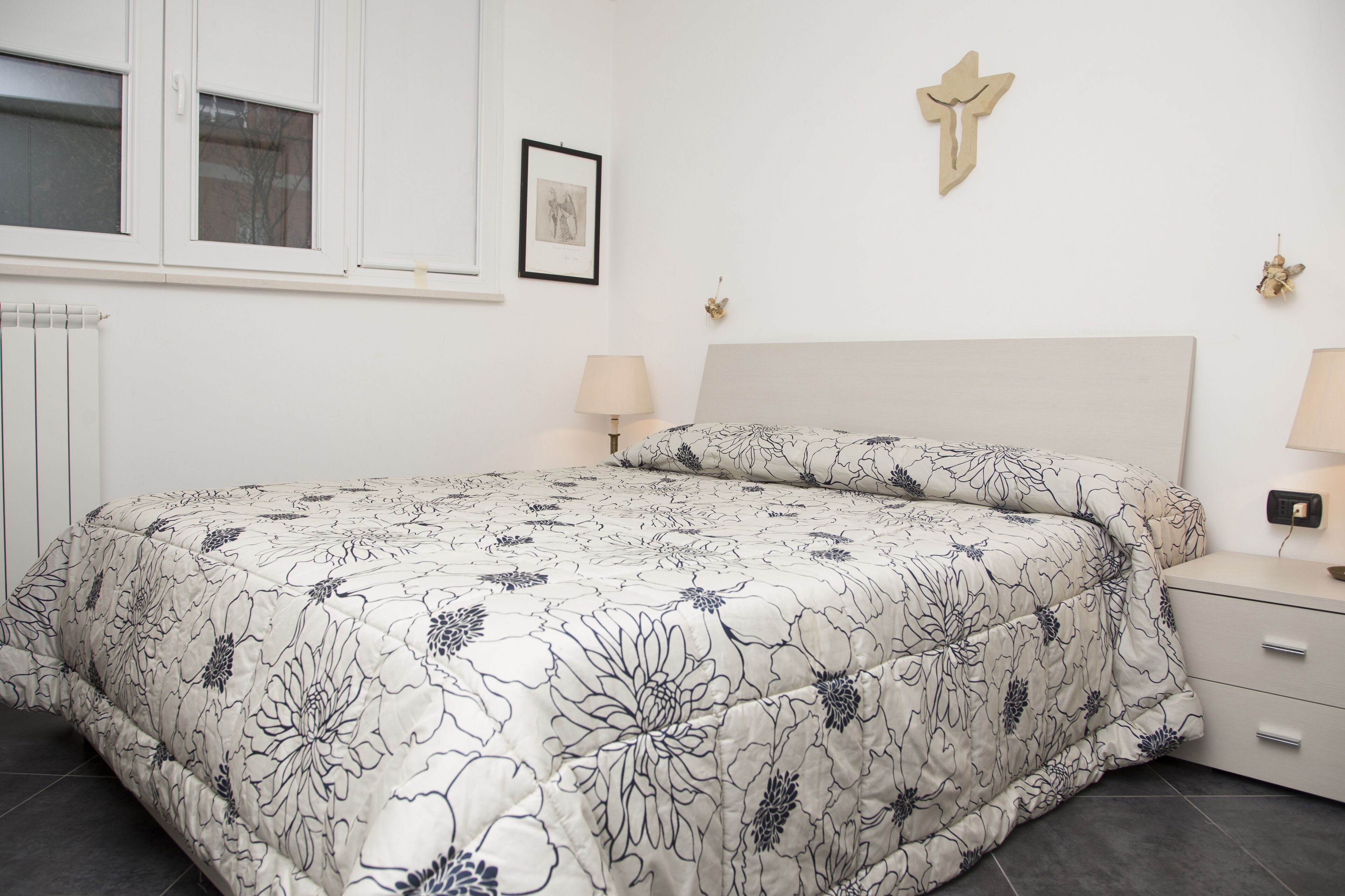 Une chambre à coucher avec une couvre-lit à motifs floraux, une tête de lit blanche, des lampes de chevet, un tableau encadré et un crucifix fixé au mur.