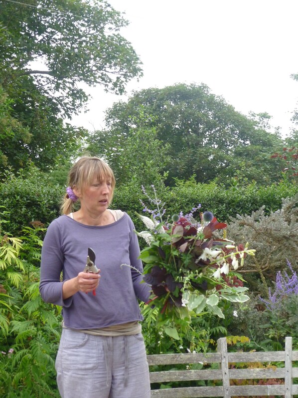A person holding a pair of pruning shears and a bouquet of flowers in a garden with dense foliage and a wooden fence. - Beautiful Historic Cottage In 540m2 Walled Garden (Helston)