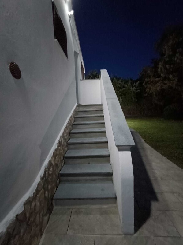 Una serie de escalones exteriores conducen a un edificio con base de piedra y paredes blancas, rodeado de follaje.