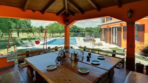 Un espace extérieur aménagé pour les repas, avec une table en bois, des chaises et une vue sur une piscine et un jardin.