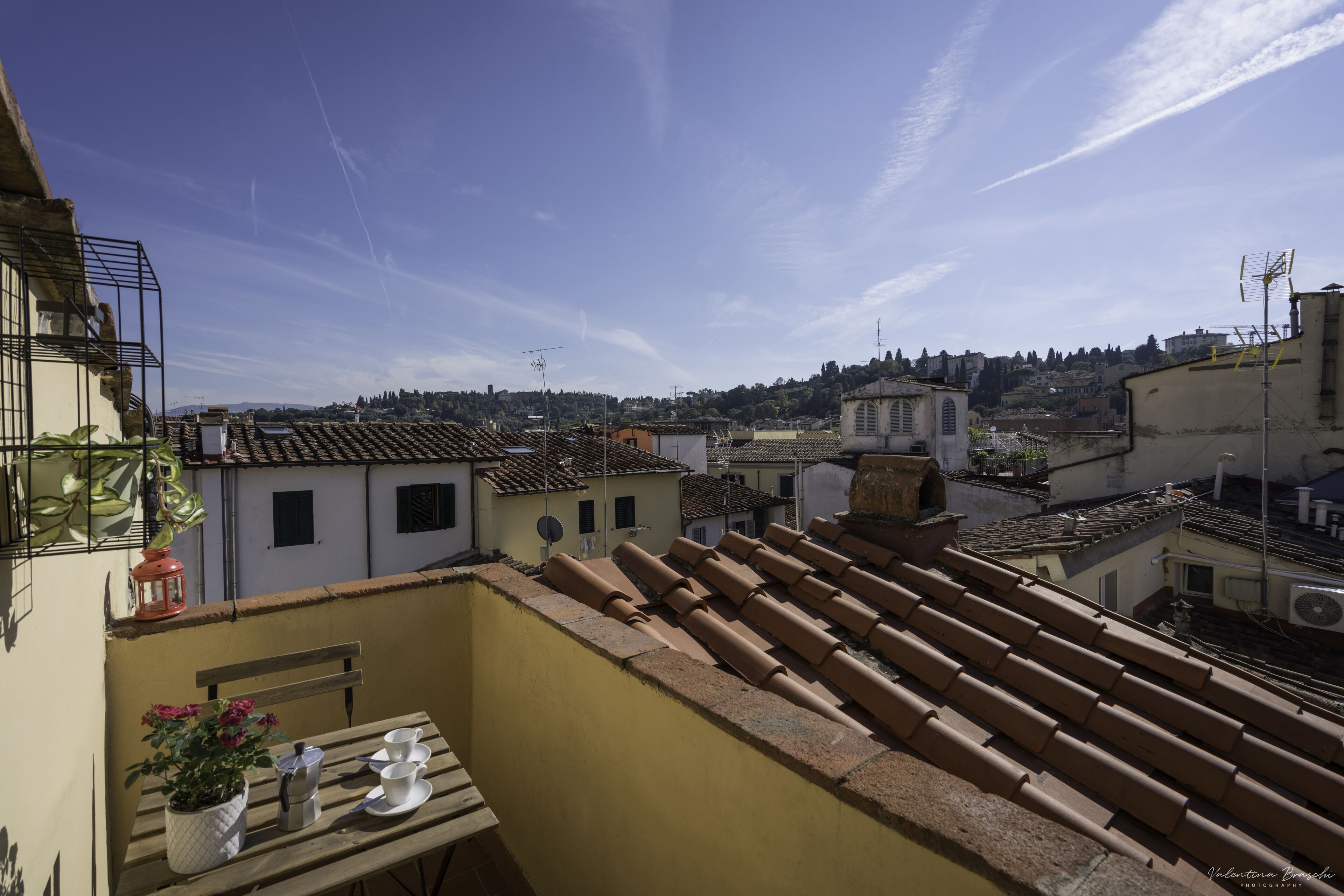 A rooftop terrace with terracotta tiles, a wooden table with a potted plant, and a view of the cityscape.