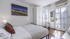 Um quarto com cama, mesa de cabeceira, luminária, janela com cortinas e uma pintura de paisagem urbana na parede.