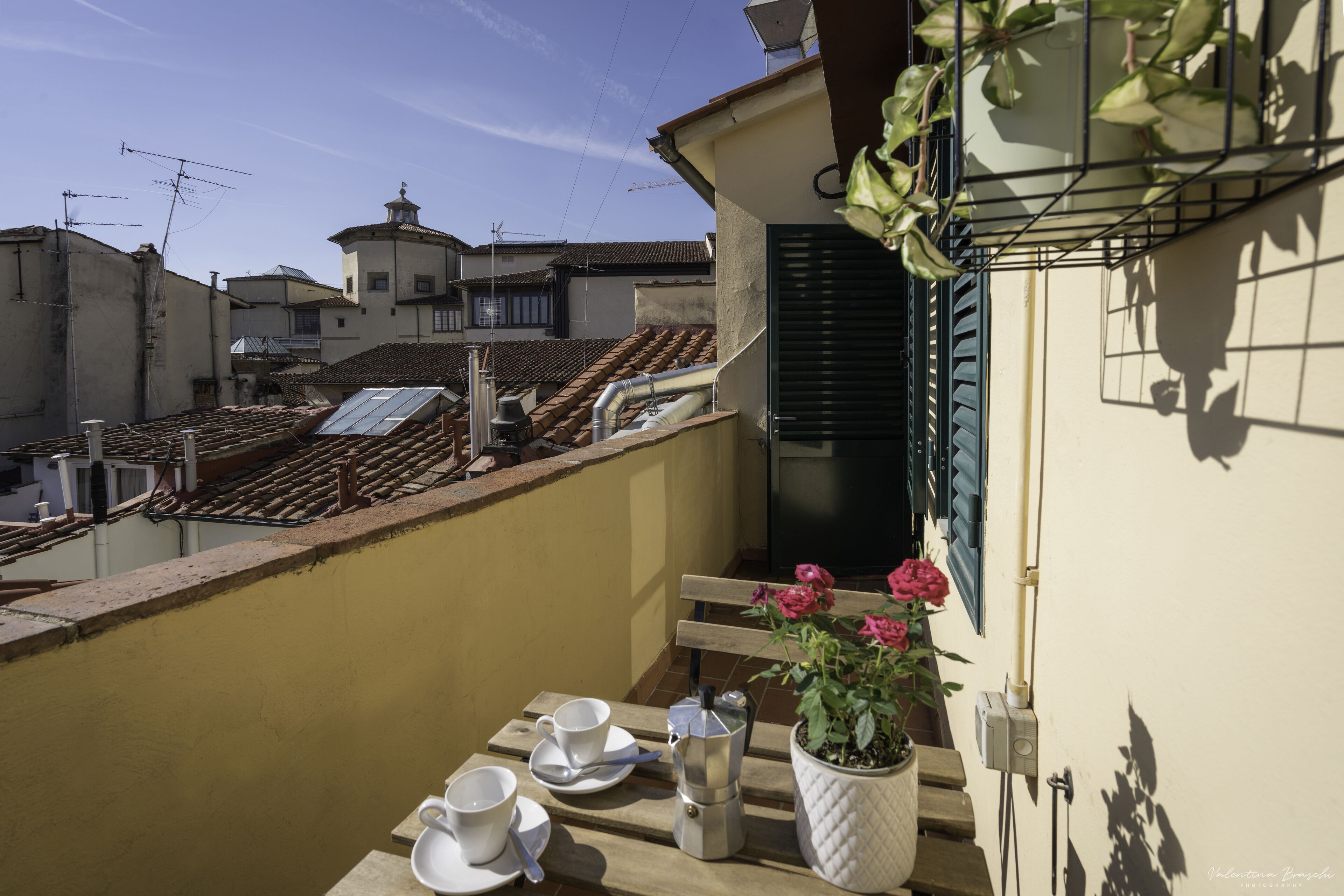 Varanda com mesa posta para dois, planta em vaso e vista para edifícios vizinhos.