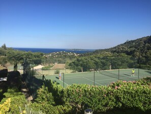 A tennis court surrounded by greenery and a view of the sea.
