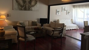 Ein Wohnzimmer mit Sofa, Sesseln, Couchtisch und Fernseher. An der Wand hängt ein Bild und ein Fenster mit Vorhängen.