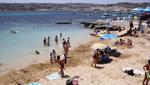 En strand med folk som solar och badar, parasoller och båtar i bakgrunden.