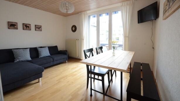Ein modernes Wohnzimmer mit einem dunklen Sofa, einem Holzesstisch und Stühlen. Ein Flachbildfernseher ist an der Wand befestigt, und es gibt drei Wanddekorationen mit Tiermotiven.