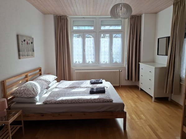 A bedroom with a wooden bed, a radiator, a window with curtains, a bedside table, and a dresser.
